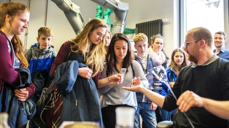Schülerinnen und Schüler schauen einem Mitarbeiter in der Glasbläserei an der Universität Jena bei der Arbeit zu.